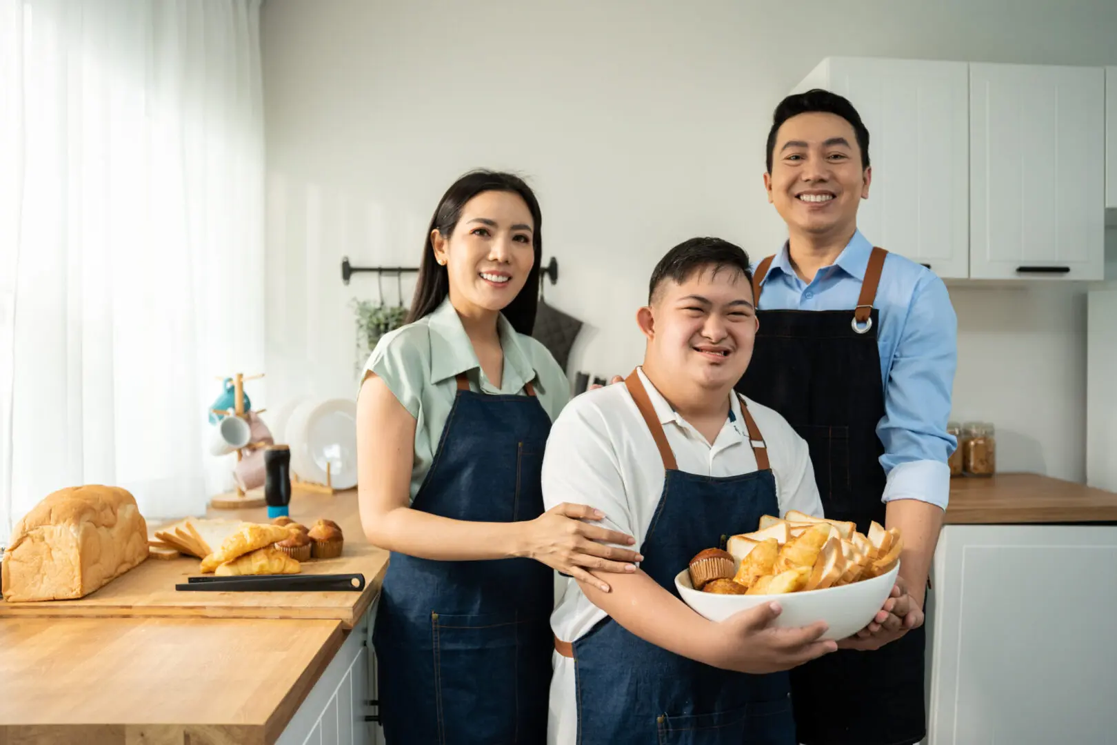 Three people in aprons smiling and holding a bowl of food in a bright kitchen.