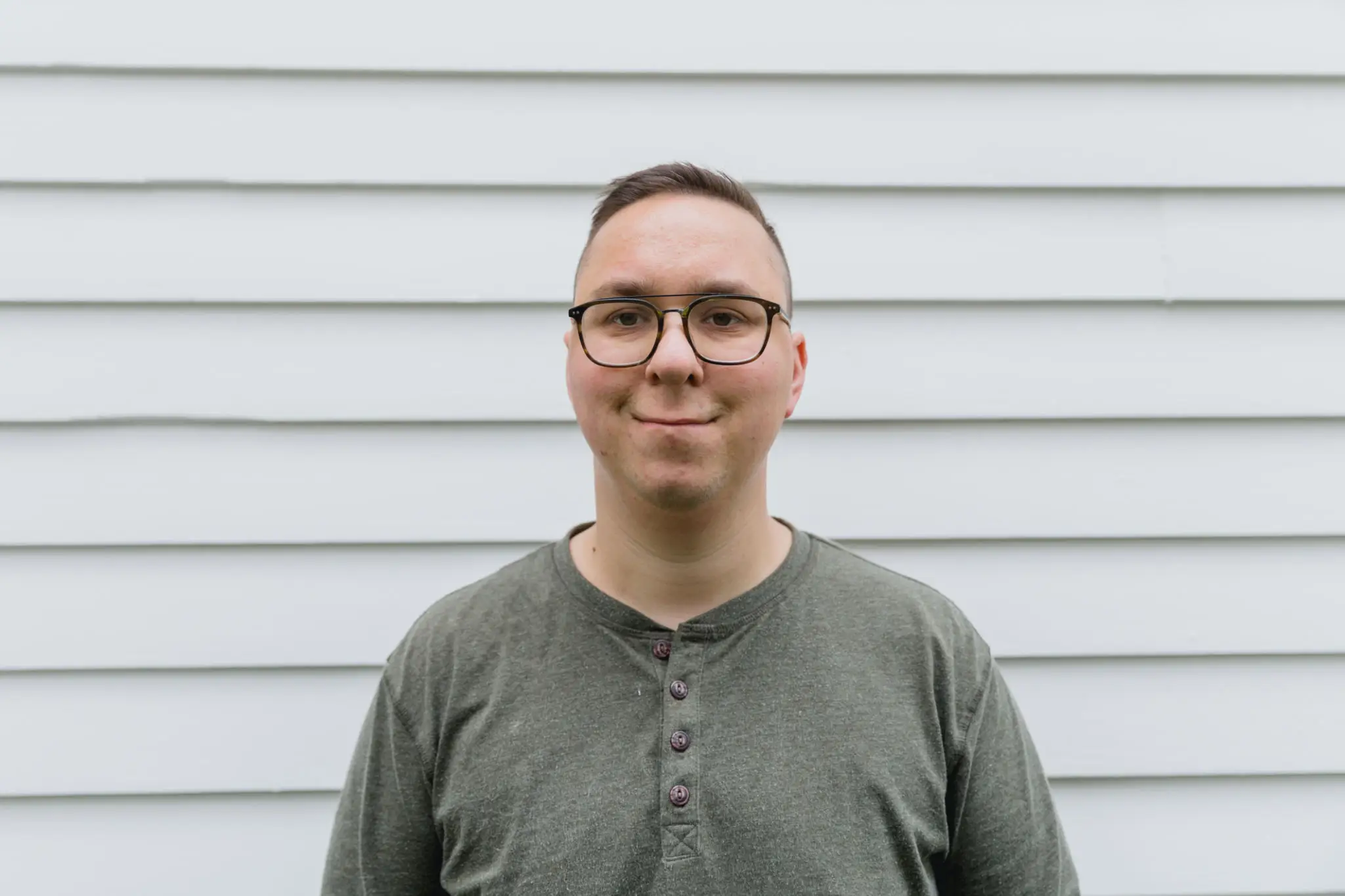 Smiling young man wearing glasses and a green shirt standing against a white wall.