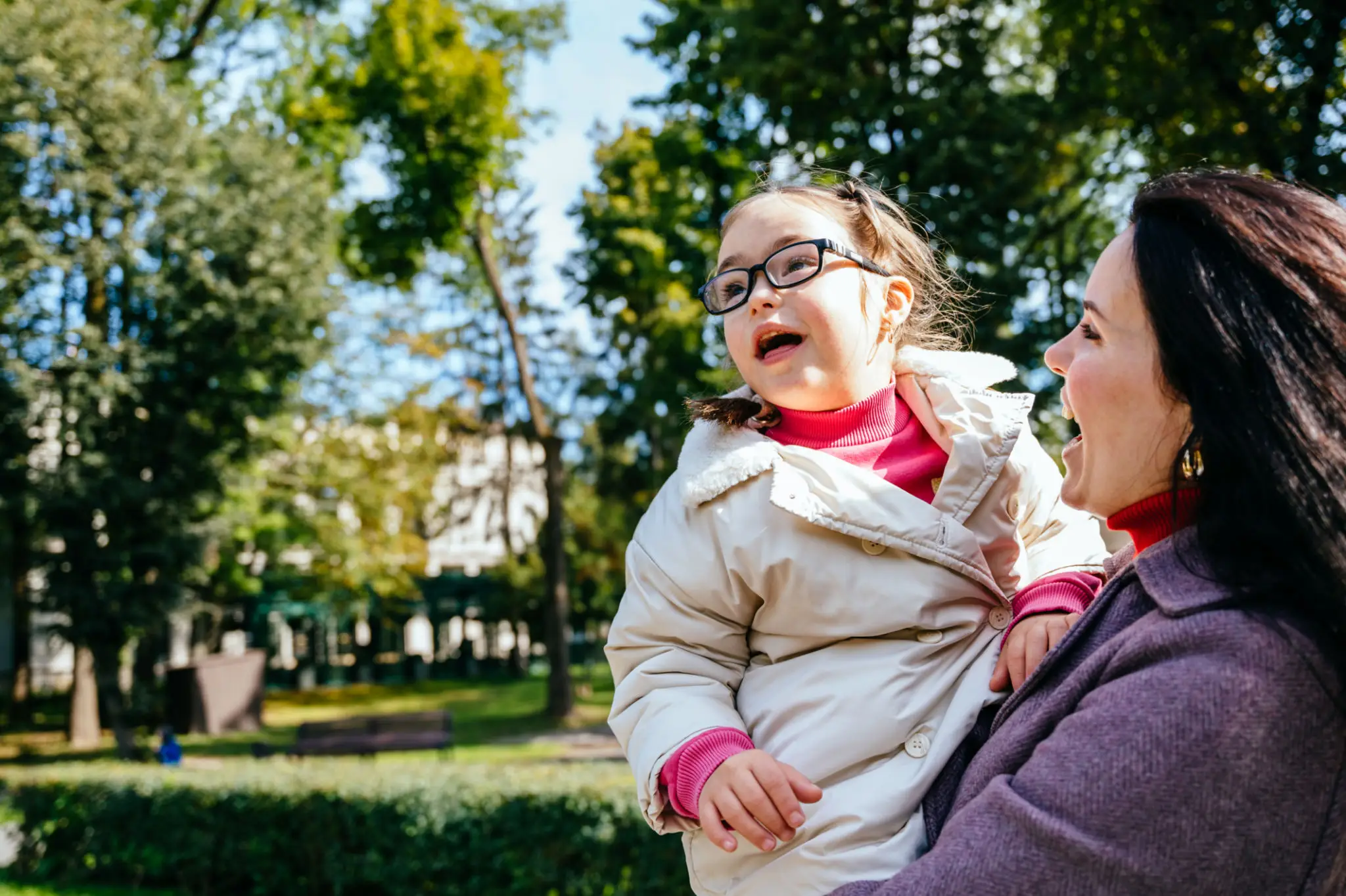 A joyful child in glasses being held by a smiling adult outdoors.